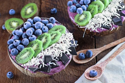 Açai smoothie bowl with fresh kiwi, frozen blueberries, organic coconut and dark chocolate pieces with wooden spoons served in coconut bowls over a rustic table. Shot from above / overhead with selective focus and blurred background.