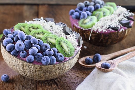 Açai smoothie bowl with fresh kiwi, frozen blueberries, organic coconut and dark chocolate pieces with wooden spoons served in coconut bowls over a rustic table. Selective focus with blurred background.