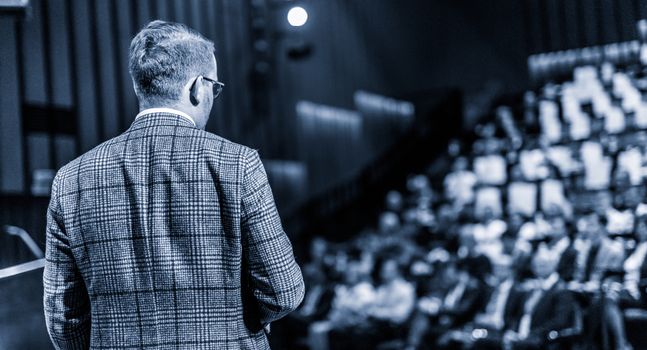 Speaker giving a talk on corporate business conference. Unrecognizable people in audience at conference hall. Business and Entrepreneurship event. Black and white, blue toned image.