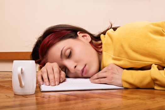 Young woman sleeping on an open notebook on the floor with a cup of coffee next to her. Beautiful young brunette student girl fell asleep on the floor preparing for an exam.