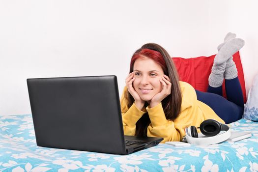 Beautiful young woman in comfortable clothes lying in bed watching something on her laptop and smiling.