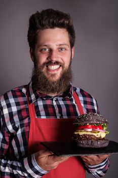 Happy smiling male cook in red apron proudly presents fresh self made black burger , gray background