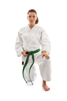 Young female martial arts fighter in white kimono and green belt kneeling on one knee and looking serious, isolated on white background. Body and spirit bent to my will.