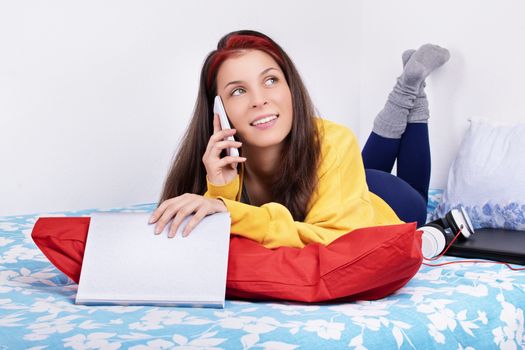 Communication, friendship concept. Beautiful young woman lying on the bed in her room with feet in the air talking on the mobile phone. Young girl on the phone talking about a good book.