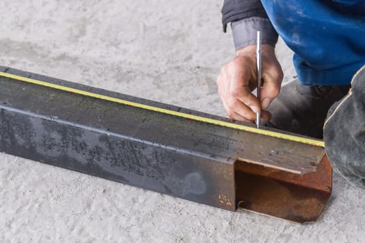 A welder writes on black huge square pipe with yellow measuring tape on concrete floor near construction place.