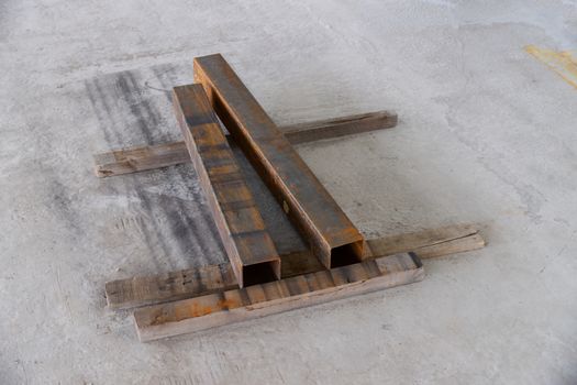 Two rusty metal square pipes on wood boards on concrete floor near construction place.