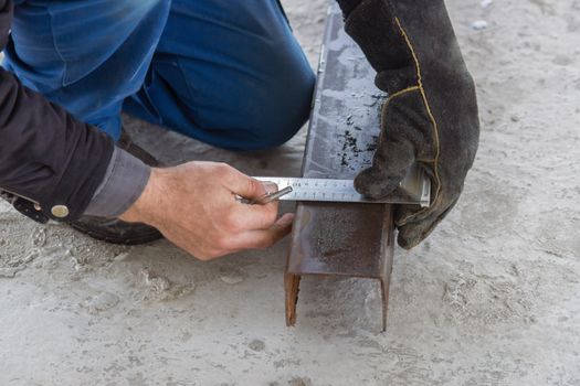 A builder draws on a square metal pipe using a setsquare on the concrete floor.