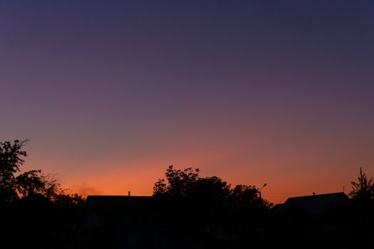 Evening sunset in the village a far away from the city. The sky is saturated with orange and blue colours. On foreground silhouttes of trees. Underexposed low light photo.