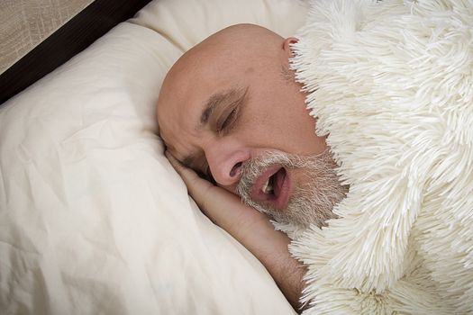 Gray-haired man sleeping on the bed under a fluffy plaid.