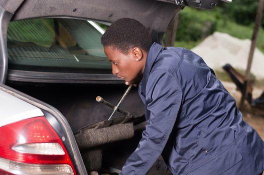 A smiling mechanic looks for her work tools in the back of a car in the garage.