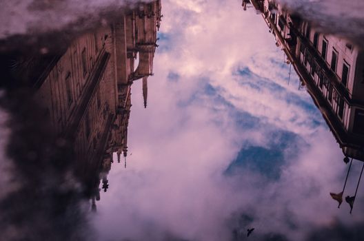 Side of Sevilla cathedral and street are reflected in a wter puddle showing the blue cloudy sky