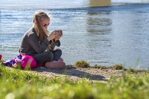 A little girl is sitting on the bank of the river with a telephone.