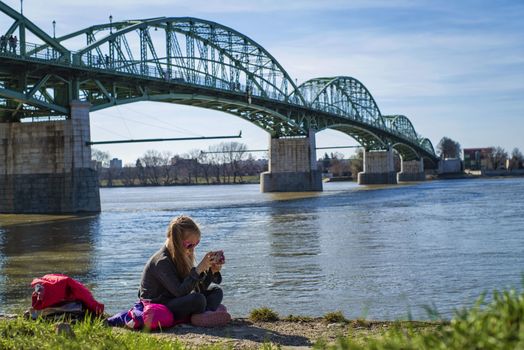 A little girl is sitting on the bank of the river with a telephone.