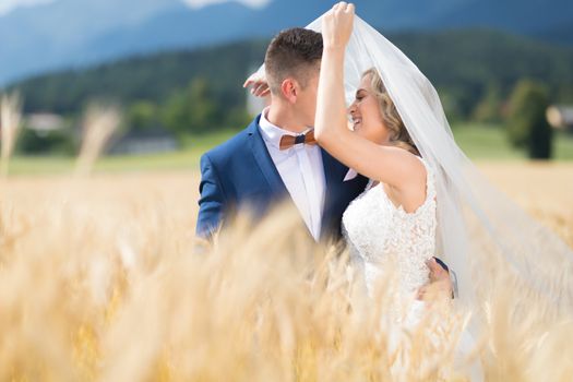 Groom hugs bride tenderly while wind blows her veil in wheat field somewhere in Slovenian countryside. Caucasian happy romantic young couple celebrating their marriage.