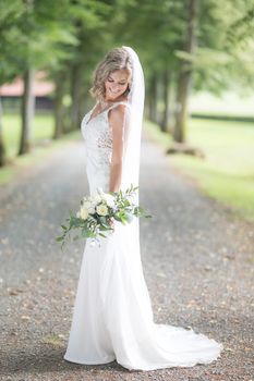 Full length portrait of beautiful sensual young blond bride in long white wedding dress and veil, holding bouquet outdoors in natural background.