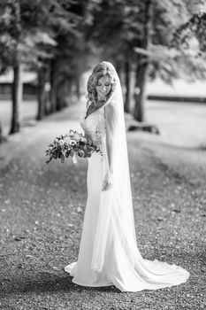 Full length portrait of beautiful sensual young blond bride in long white wedding dress and veil, holding bouquet outdoors in natural background. Black and white photo.