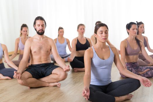 Group of young sporty attractive people in yoga studio, practicing yoga lesson with instructor, sitting on floor in Siddhasana, easy seated yoga pose. Healthy active lifestyle, working out in gym.