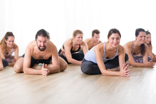 Group of young sporty attractive people in yoga studio, practicing yoga lesson with instructor, sitting on floor in forward band stretching yoga pose. Healthy active lifestyle, working out in gym.