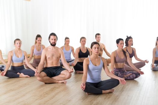 Group of young sporty attractive people in yoga studio, practicing yoga lesson with instructor, sitting on floor in Siddhasana, easy seated yoga pose. Healthy active lifestyle, working out in gym.