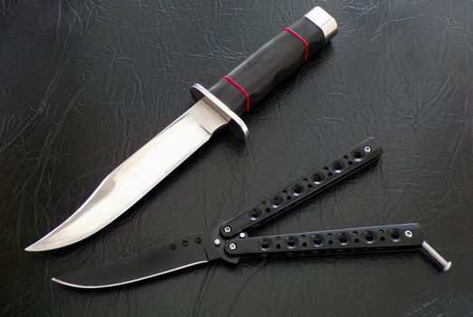 Knives close-up. Weapons for murderers and maniacs. Knives on a background of black leather. Butterfly knife and Bowie hunting knife.