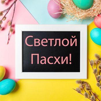 Orthodox Easter concept. Colorful eggs and pussy willow branches on bright colorful background and chalkboard with russian letters Happy Easter. Top down view or flat lay. Square crop for social media