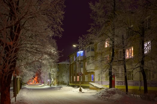 Night snowy street with trees in the light of lanterns and windows of houses painted with different colors.