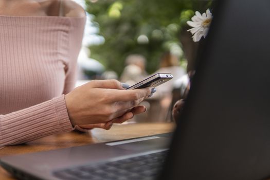 Modern business woman is reading incoming sms message on smartphone or watching broadcasting online on modern mobile phone while sitting in caffe with a laptop. Female student sitting in coffee