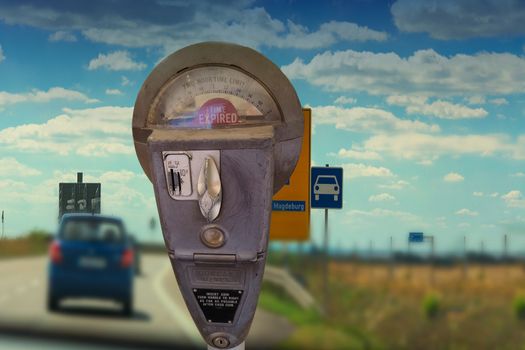 Old-fashioned parking meter with parking time display in the blurred background a road with a car in front of blue sky.