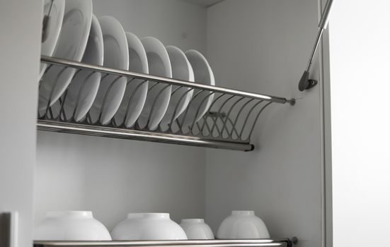 Dish drying metal rack with big nice white clean plates. Traditional comfortable kitchen. Open white dish draining closet with wet dishes of glass and ceramic, plates, bowls drying inside on rack