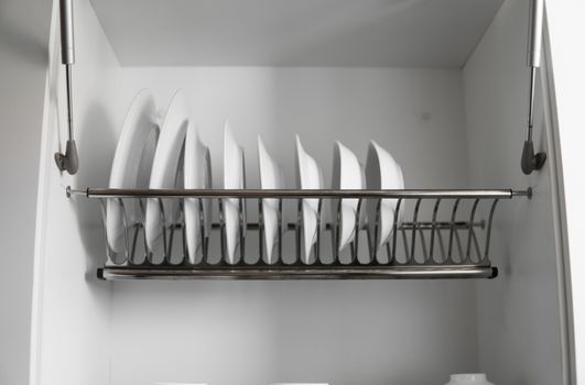 Dish drying metal rack with big nice white clean plates. Traditional comfortable kitchen. Open white dish draining closet with wet dishes of glass and ceramic, plates, bowls drying inside on rack