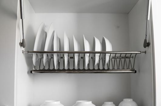 Dish drying metal rack with big nice white clean plates. Traditional comfortable kitchen. Open white dish draining closet with wet dishes of glass and ceramic, plates, bowls drying inside on rack