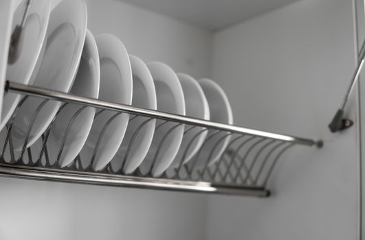 Dish drying metal rack with big nice white clean plates. Traditional comfortable kitchen. Open white dish draining closet with wet dishes of glass and ceramic, plates, bowls drying inside on rack
