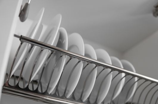 Dish drying metal rack with big nice white clean plates. Traditional comfortable kitchen. Open white dish draining closet with wet dishes of glass and ceramic, plates, bowls drying inside on rack