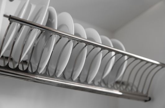 Dish drying metal rack with big nice white clean plates. Traditional comfortable kitchen. Open white dish draining closet with wet dishes of glass and ceramic, plates, bowls drying inside on rack