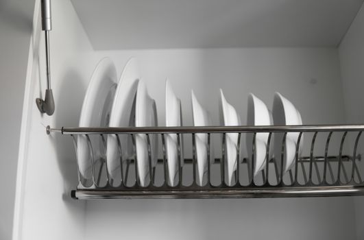Dish drying metal rack with big nice white clean plates. Traditional comfortable kitchen. Open white dish draining closet with wet dishes of glass and ceramic, plates, bowls drying inside on rack