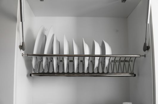 Dish drying metal rack with big nice white clean plates. Traditional comfortable kitchen. Open white dish draining closet with wet dishes of glass and ceramic, plates, bowls drying inside on rack