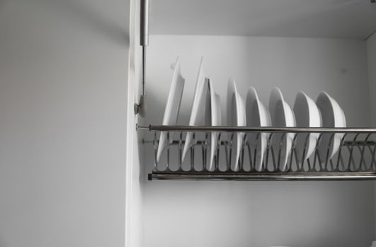 Dish drying metal rack with big nice white clean plates. Traditional comfortable kitchen. Open white dish draining closet with wet dishes of glass and ceramic, plates, bowls drying inside on rack