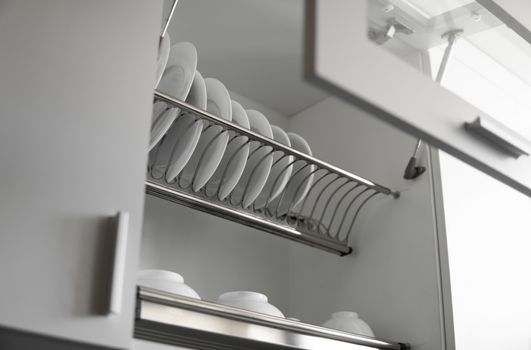 Dish drying metal rack with big nice white clean plates. Traditional comfortable kitchen. Open white dish draining closet with wet dishes of glass and ceramic, plates, bowls drying inside on rack