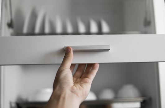 Hand holds the door of the dryer with utensils. Dish drying metal rack with white clean plates. Traditional comfortable kitchen. Open white dish draining closet with wet dishes of plates, bowls