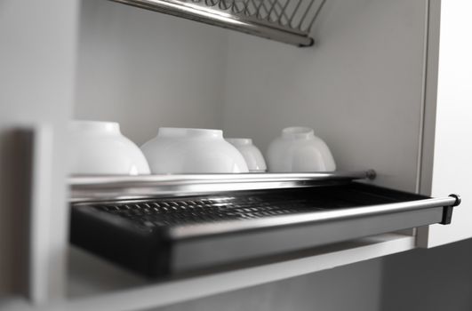 Dish drying metal rack with big nice white clean plates. Traditional comfortable kitchen. Open white dish draining closet with wet dishes of glass and ceramic, plates, bowls drying inside on rack