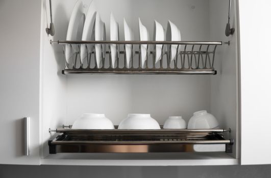 Dish drying metal rack with big nice white clean plates. Traditional comfortable kitchen. Open white dish draining closet with wet dishes of glass and ceramic, plates, bowls drying inside on rack