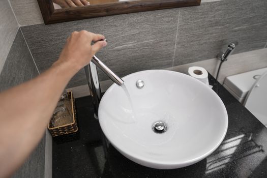 Male hand use a faucet in a bathroom interior with white round sink and chrome faucet. Water flowing from the chrome faucet