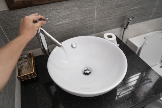 Male hand use a faucet in a bathroom interior with white round sink and chrome faucet. Water flowing from the chrome faucet