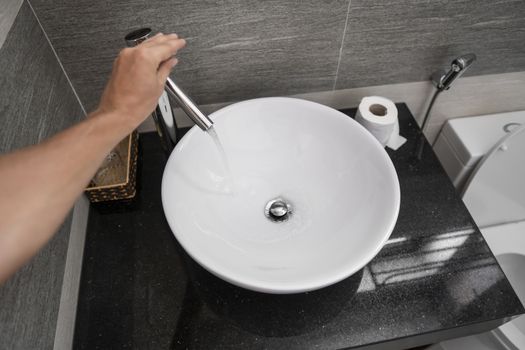 Male hand use a faucet in a bathroom interior with white round sink and chrome faucet. Water flowing from the chrome faucet