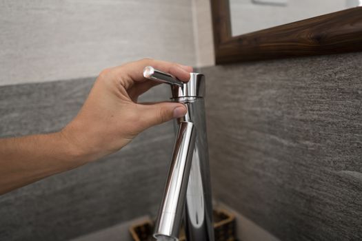 Male hand use a faucet in a bathroom interior with white round sink and chrome faucet. Water flowing from the chrome faucet