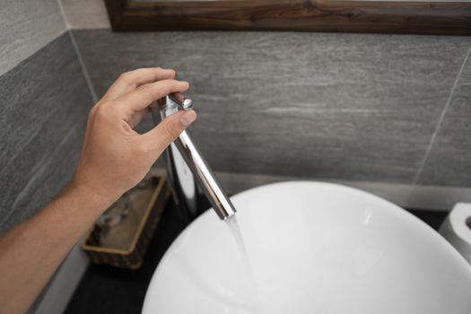 Male hand use a faucet in a bathroom interior with white round sink and chrome faucet. Water flowing from the chrome faucet