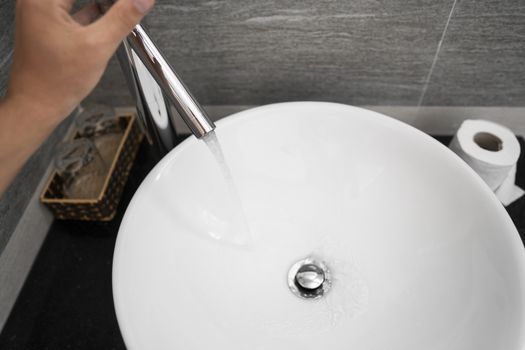 Male hand use a faucet in a bathroom interior with white round sink and chrome faucet. Water flowing from the chrome faucet