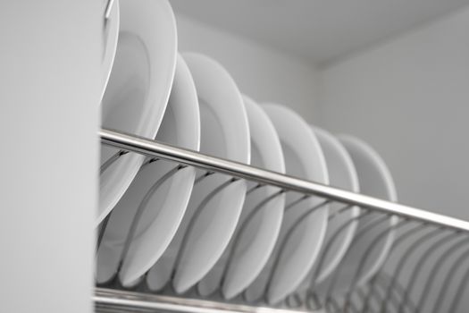 Dish drying metal rack with big nice white clean plates. Traditional comfortable kitchen. Open white dish draining closet with wet dishes of glass and ceramic, plates, bowls drying inside on rack