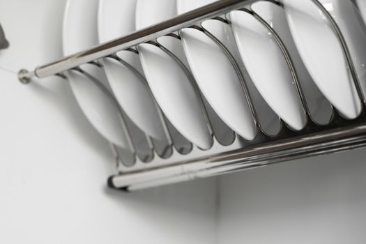 Dish drying metal rack with big nice white clean plates. Traditional comfortable kitchen. Open white dish draining closet with wet dishes of glass and ceramic, plates, bowls drying inside on rack