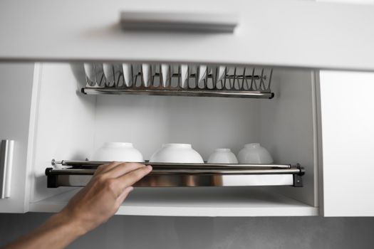 Dish drying metal rack with big nice white clean plates. Traditional comfortable kitchen. Open white dish draining closet with wet dishes of glass and ceramic, plates, bowls drying inside on rack